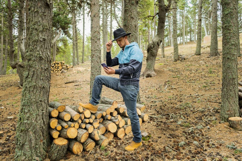 Man black cowboy hat taking a selfie on cell phone on pile wood logs in beautiful forest landscape.Male using smartphone in nature
