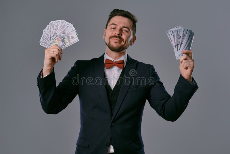 Man in black suit and red bow-tie is showing two fans of hundred dollar bills, posing on gray studio background stock photography