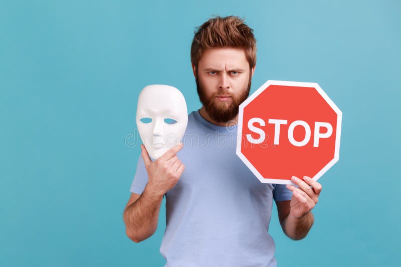 Portrait of bossy serious handsome bearded man holding red stop sign and white mask, looking at camera with strict expression. Indoor studio shot isolated on blue background. Portrait of bossy serious handsome bearded man holding red stop sign and white mask, looking at camera with strict expression. Indoor studio shot isolated on blue background.