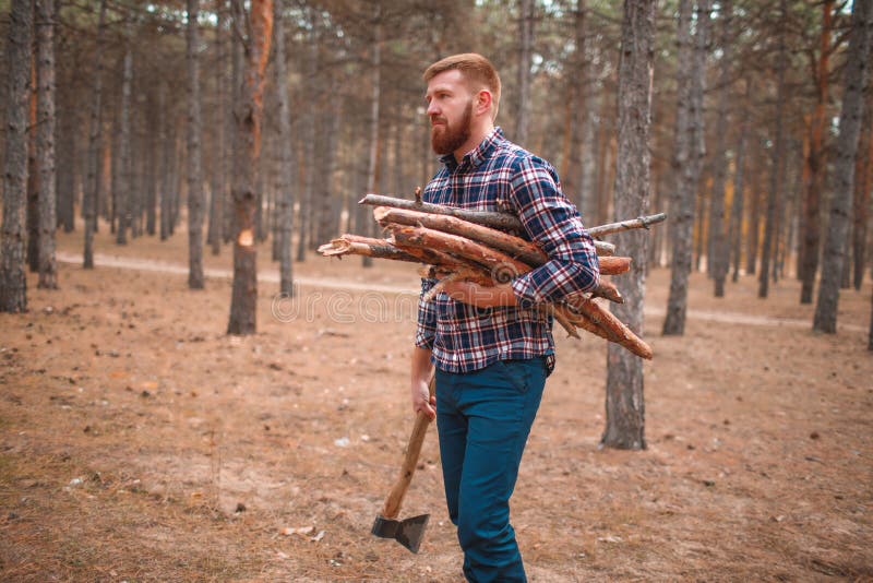 A man in a checkered shirt, carries a bunch of firewood and an ax in the forest. Outdoors.