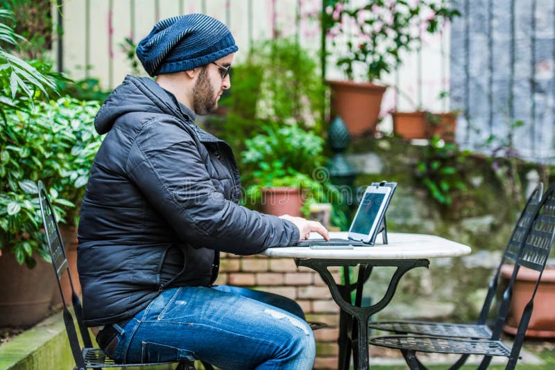 Muž na sobě kabát a klobouk, sedící v zahradě a kontrola sociálních médií na jeho tabletu.