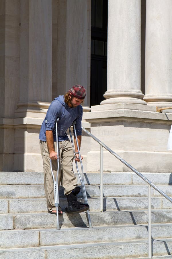 Man on crutches and wearing an ankle cast makes his way down concrete steps in front of a county courthouse.
