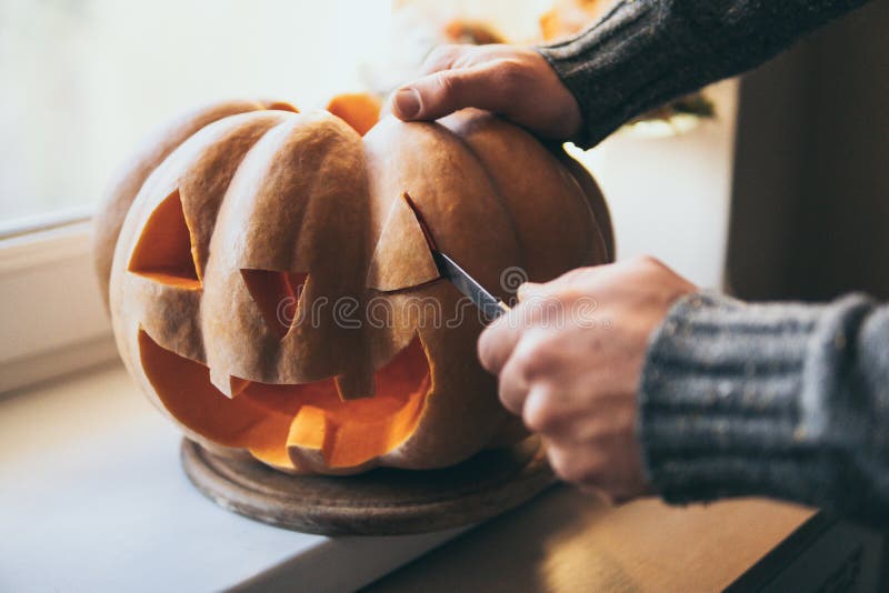Man cutting pumpkin for Halloween party with a small knife, closeup on hands, carve, decoration, big, head, orange, vegetable, scary, face, smile, eye, mouth, open, tradition, holiday, celebration, evening, kitchen, make, saints, thanksgiving, copy, space, masculine, strong, sweater, autumn, fall, harvest, crop, home, windowsill, spooky, fear, vegan, fun, smiling, allhalloween, christian, trick-or-treat, horror, vigil. Man cutting pumpkin for Halloween party with a small knife, closeup on hands, carve, decoration, big, head, orange, vegetable, scary, face, smile, eye, mouth, open, tradition, holiday, celebration, evening, kitchen, make, saints, thanksgiving, copy, space, masculine, strong, sweater, autumn, fall, harvest, crop, home, windowsill, spooky, fear, vegan, fun, smiling, allhalloween, christian, trick-or-treat, horror, vigil