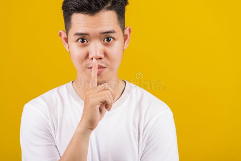 Asian handsome young man holding index finger on lips asking for silence or secret gesturing, studio shot isolated on yellow background, making shh shush gesture concept. Asian handsome young man holding index finger on lips asking for silence or secret gesturing, studio shot isolated on yellow background, making shh shush gesture concept