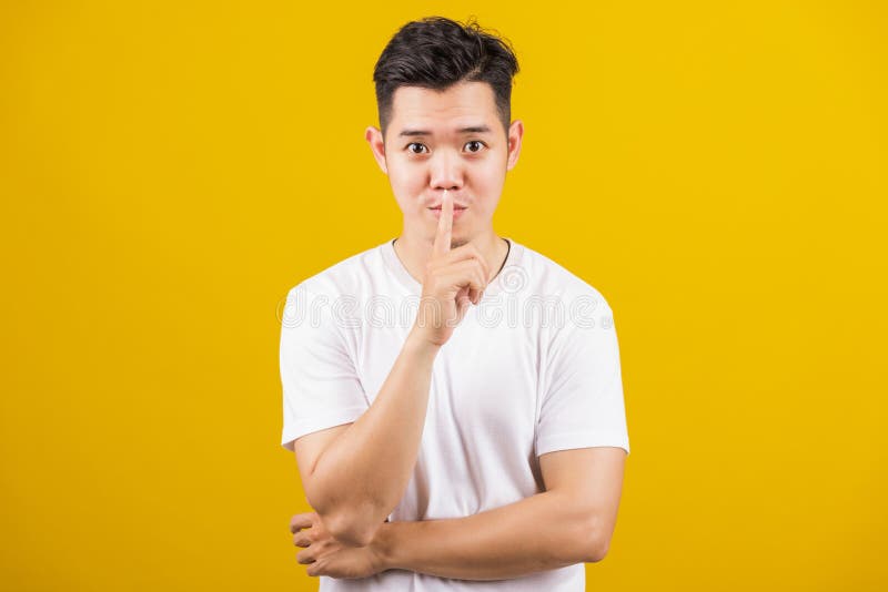 Asian handsome young man holding index finger on lips asking for silence or secret gesturing, studio shot isolated on yellow background, making shh shush gesture concept. Asian handsome young man holding index finger on lips asking for silence or secret gesturing, studio shot isolated on yellow background, making shh shush gesture concept