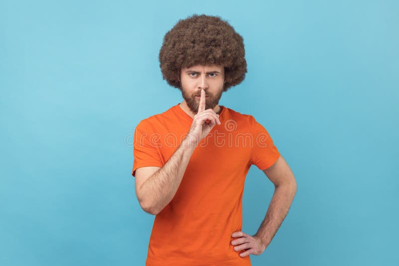 Portrait of man with Afro hairstyle wearing orange T-shirt holding finger on lips making hush silence gesture, asking to keep secret, don&#x27;t speak. Indoor studio shot isolated on blue background. Portrait of man with Afro hairstyle wearing orange T-shirt holding finger on lips making hush silence gesture, asking to keep secret, don&#x27;t speak. Indoor studio shot isolated on blue background.