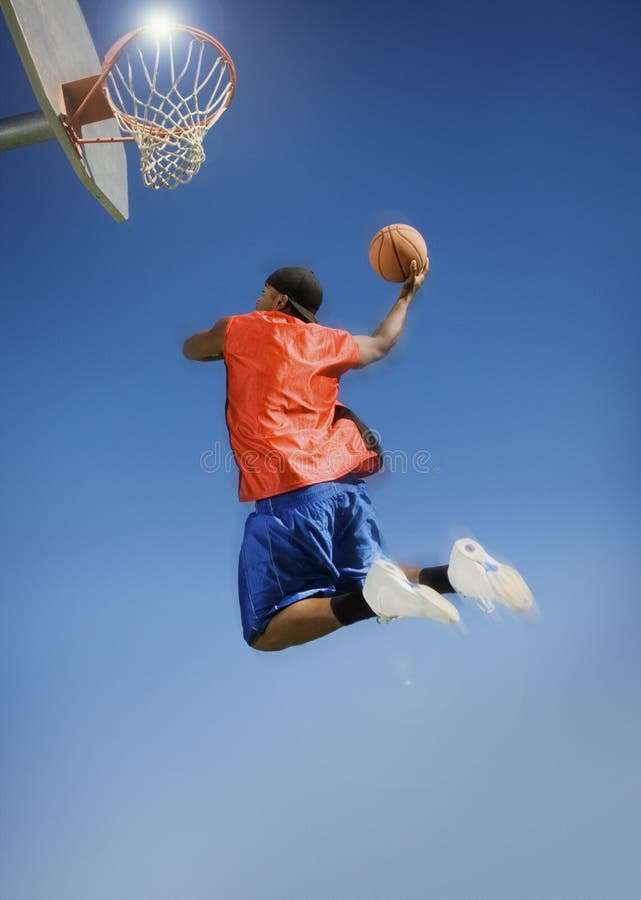 Low angle view of man dunking basketball into hoop against clear blue sky. Low angle view of man dunking basketball into hoop against clear blue sky