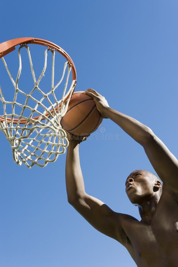 Low angle view of determined young man dunking basketball into hoop against clear blue sky. Low angle view of determined young man dunking basketball into hoop against clear blue sky