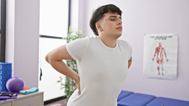 Man experiencing back pain in a physical therapy clinic with exercise equipment and anatomy poster royalty free stock image