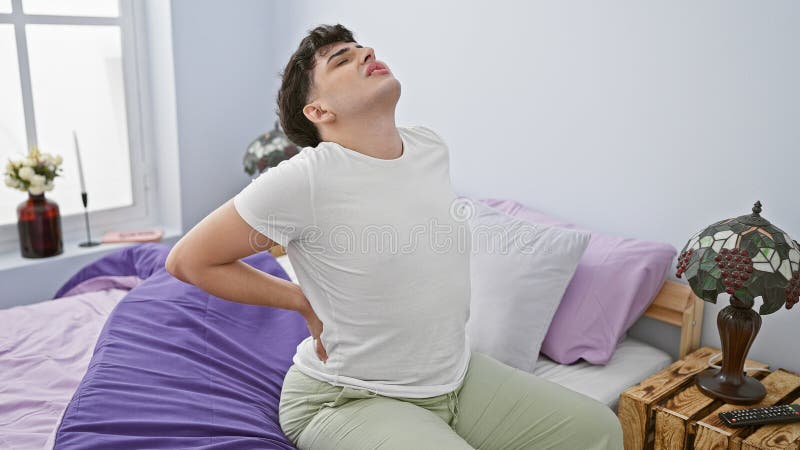 A man expressing back pain while sitting on the bed in a modern bedroom interior setting stock photos