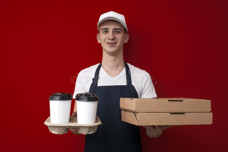 Young guy a food delivery uniform in uniform gives coffee and pizza boxes on a red background, food delivery service worker. Young guy a food delivery uniform in uniform gives coffee and pizza boxes on a red background, food delivery service worker