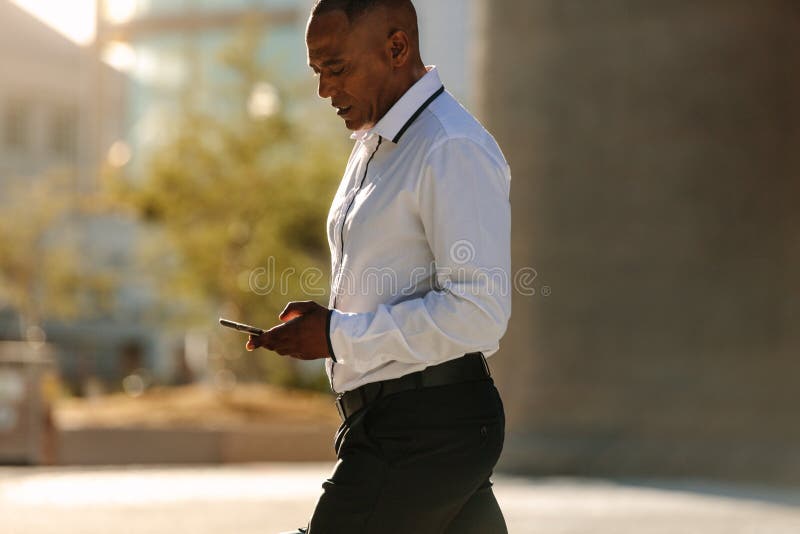 Man in formal clothes looking at his mobile phone while walking on street. Busy office going person walking on street and using his cellphone. Man in formal clothes looking at his mobile phone while walking on street. Busy office going person walking on street and using his cellphone.