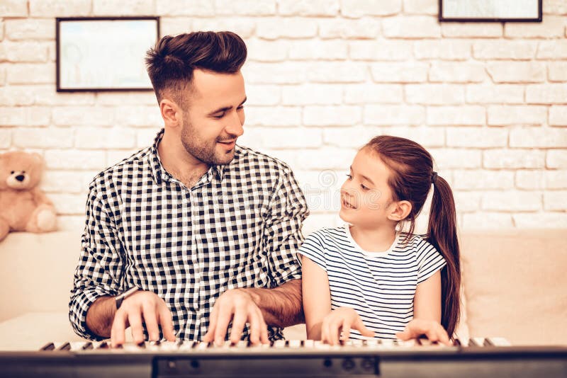 Man and Girl Play Piano. Play Piano Together. Father and Daughter on Sofa. Spend Time Together. Father`s day. Man Play with Girl. White Interior. Girl wiht Man on Sofa. Play Piano and Sing