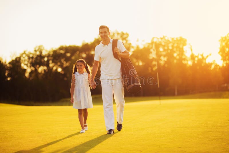 The man and the girl are walking along the field for the yagolph holding hands. A man has a bag with golf clubs