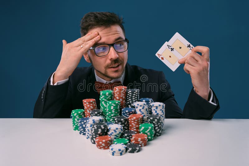 Man in glasses, black suit sitting at white table with stacks of chips, holding two playing cards, posing on blue royalty free stock photography