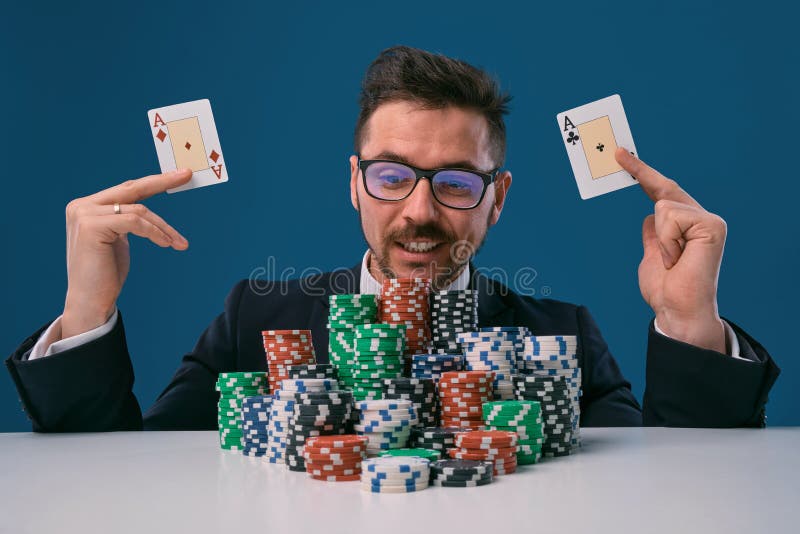 Man in glasses, black suit sitting at white table with stacks of chips, holding two playing cards, posing on blue stock photos