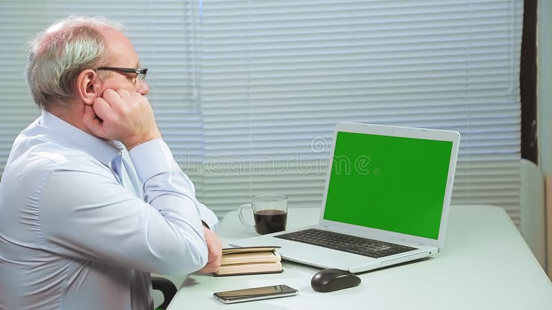 A man in glasses in an office with blinds at a computer works on a green screen and drinks coffee