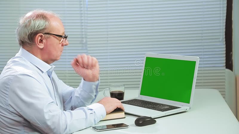 A man in glasses in an office with blinds at a computer works on a green screen and drinks coffee