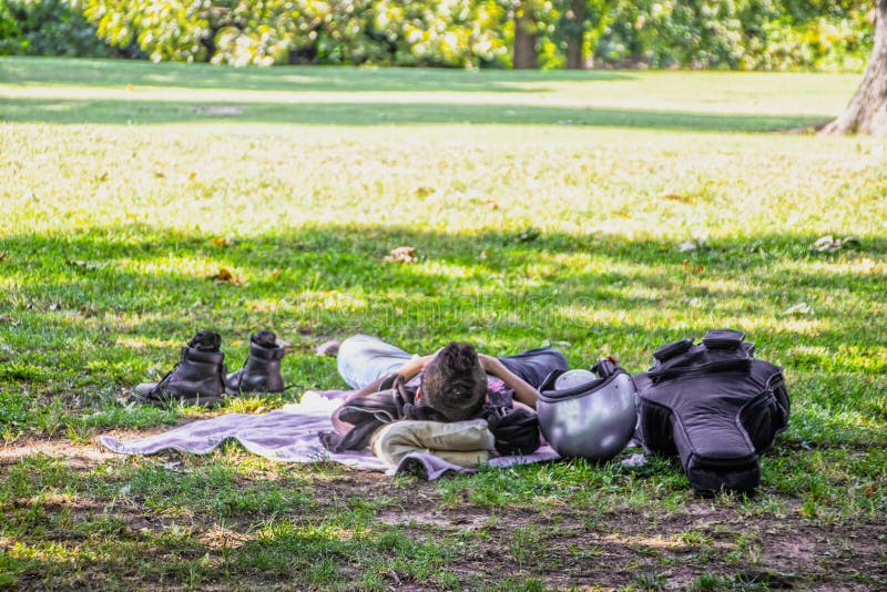 Man with guitar case and motorcycle helment with boots off lying in shade napping under tree in leafy park - Room for copy.