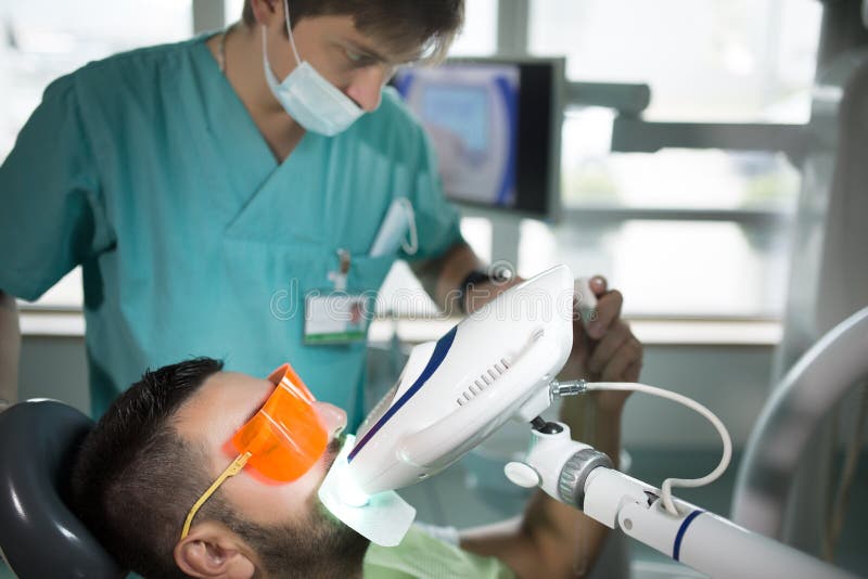 Man having teeth whitening by dental UV whitening device,dental assistant taking care of patient.Eyes protected with glasses royalty free stock image Man having teeth whitening by dental UV whitening device,dental assistant taking care of patient.Eyes protected with glasses royalty free stock image