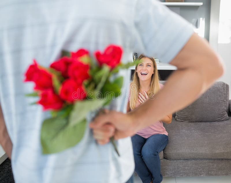 Man hiding bouquet of roses from smiling girlfriend on the couch in sitting room at home