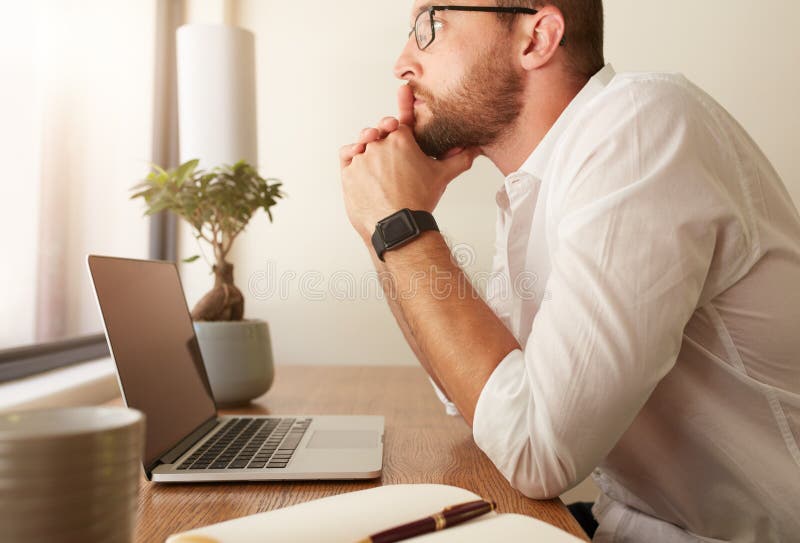 Side view of businessman sitting at his desk with a laptop and looking away. Man at his workdesk thinking of business solutions.