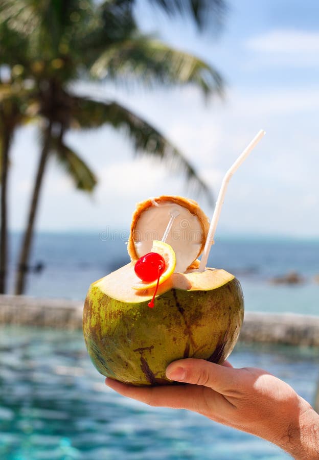 Man holding coconut cocktail with drinking straw and cocktail cherry by the swimming pool