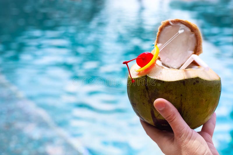 Man holding coconut cocktail with green drinking straw and cocktail cherry by the swimming pool