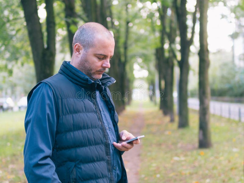 Man is holding a phone, walking in the city Park, among the trees and looking at the smartphone.