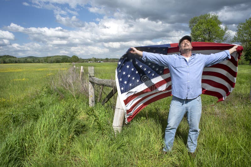 Man holding spread out American flag over behind him, standing in grass farm field looking up at the sun, barbwire fence