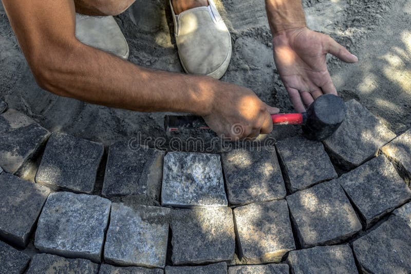 A man holds a red-black rubber mallet in his hand and knocks it on the surface of gray granite stones on the sidewalk, top view. stock photo