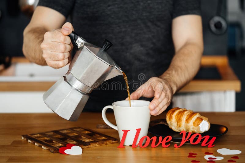 Man in home clothes pours hot coffee into a mug with the words I LOVE YOU from red paper on the table with a cake, against the bac stock photography Man in home clothes pours hot coffee into a mug with the words I LOVE YOU from red paper on the table with a cake, against the bac stock photography