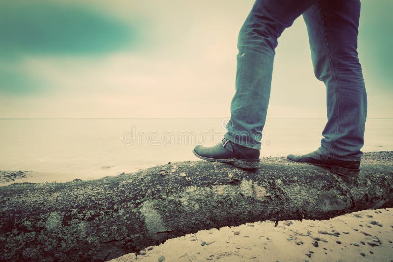 Man in jeans and elegant shoes standing on fallen tree on wild beach looking at sea. Vintage