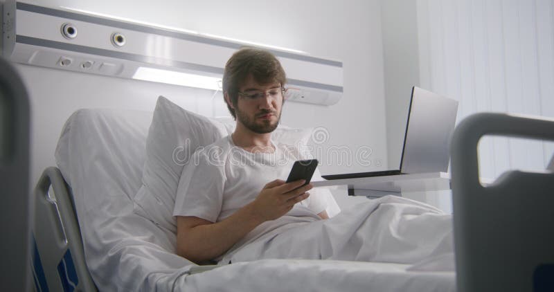 Dospělý muž spočívá na postel v nemocnice hlídat a chatování zákazník telefon.