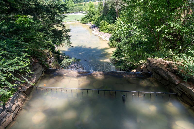 A man-made waterfall flows into a tranquil stream in a lush summer garden. The water is surrounded by green trees and a stone retaining wall. Man made waterfall stock images, royalty-free photos and pictures