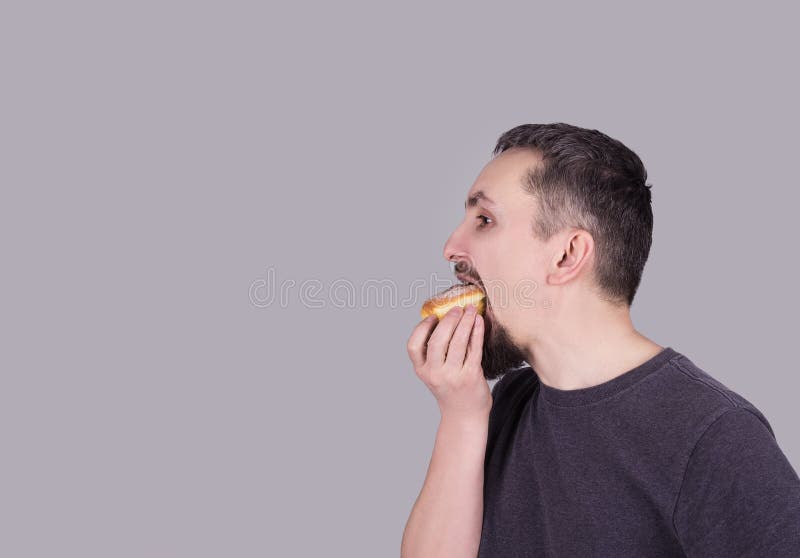 Man med ett skägg som äter en bulle över grå bakgrund arkivfoton Man med ett skägg som äter en bulle över grå bakgrund arkivfoton