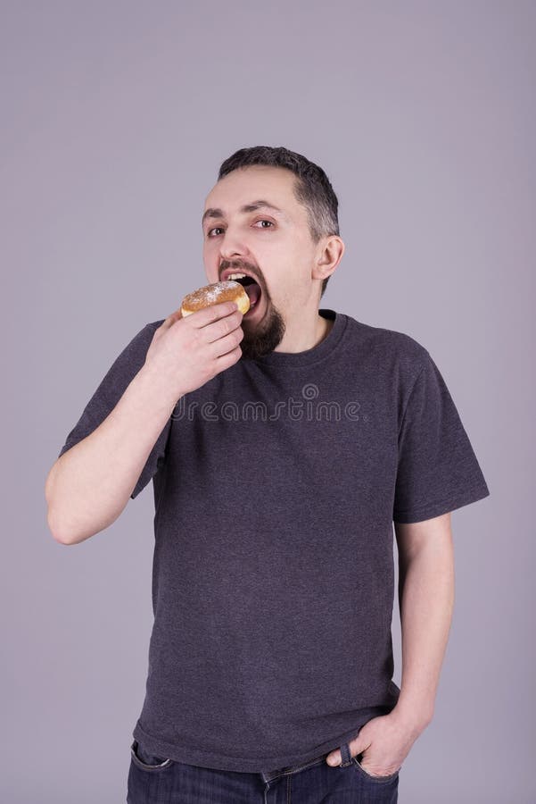 Man med ett skägg som äter en bulle över grå bakgrund royaltyfri bild Man med ett skägg som äter en bulle över grå bakgrund royaltyfri bild