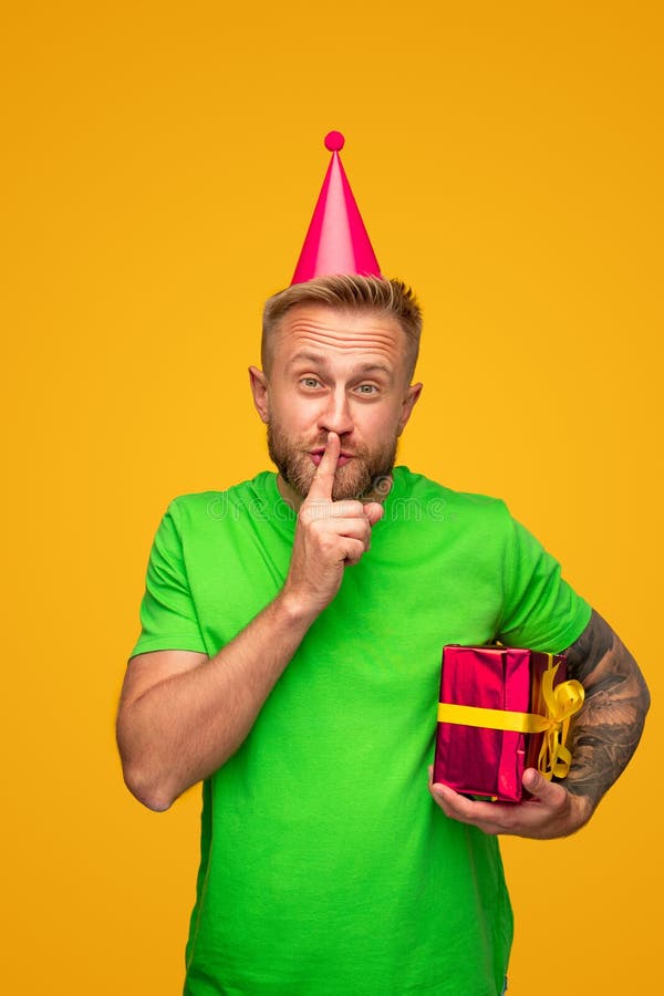 Funny adult bearded hipster guy in bright red shirt and party hat holding wrapped present box and doing shh sign against yellow background. Funny adult bearded hipster guy in bright red shirt and party hat holding wrapped present box and doing shh sign against yellow background
