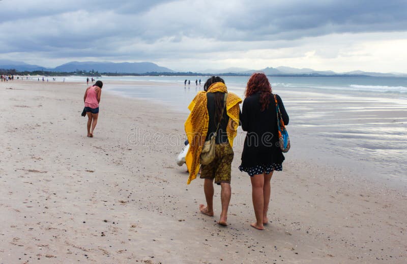 Man met grote sjaal en dreadlocks en vrouw in kleding en lange rode haargang langs strand in Augustus 2014 van Byron Bay NSW Aust stock foto Man met grote sjaal en dreadlocks en vrouw in kleding en lange rode haargang langs strand in Augustus 2014 van Byron Bay NSW Aust stock foto