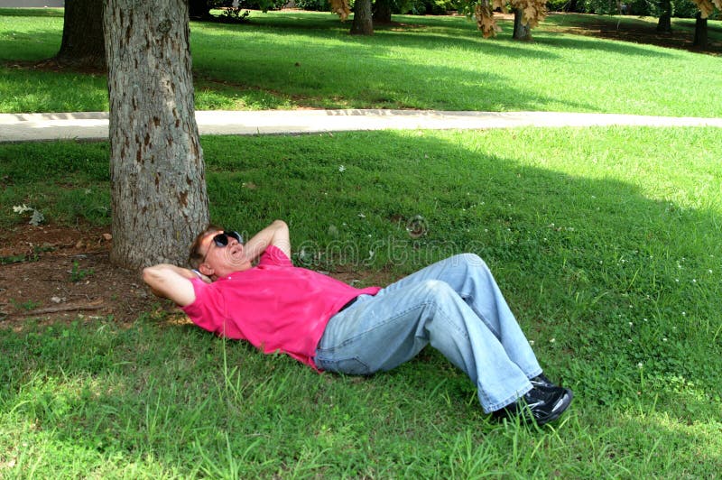 Man wearing jeans and red shirt napping in the shade under a tree. Man wearing jeans and red shirt napping in the shade under a tree