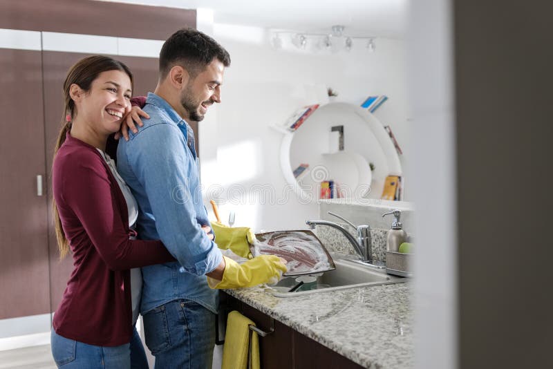 Young couple doing housework and chores. Man cleaning dishes in kitchen and girlfriend hugging him from behind. Young couple doing housework and chores. Man cleaning dishes in kitchen and girlfriend hugging him from behind