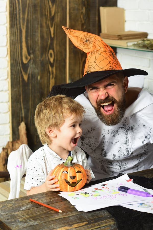 Man in orange hat and kid with happy faces. Hold carved pumpkin and draw. Witcher and little magician decorate room for Halloween. Man and boy on wooden royalty free stock photography Man in orange hat and kid with happy faces. Hold carved pumpkin and draw. Witcher and little magician decorate room for Halloween. Man and boy on wooden royalty free stock photography