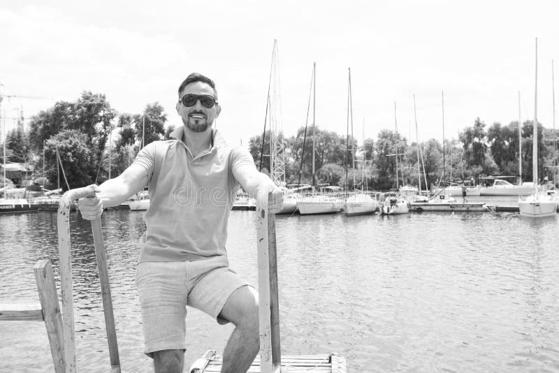Man in polo and sunglasses laddering up on boat. Happy businessman on vacation with yachts on dock and wooden pier background stock images Man in polo and sunglasses laddering up on boat. Happy businessman on vacation with yachts on dock and wooden pier background stock images