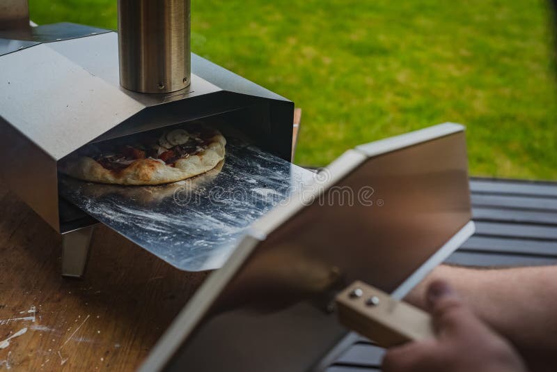 Portable wood fired pizza oven. Man is pulling a delicious fresh home made pizza out of a stainless steel home portble oven fueled by pellets. Outdoor pizza