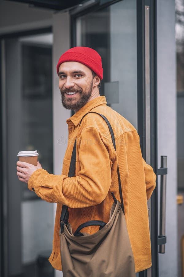 Man in a Red Hat and Orange Jacket with a Coffee Cup in Hands Looking
