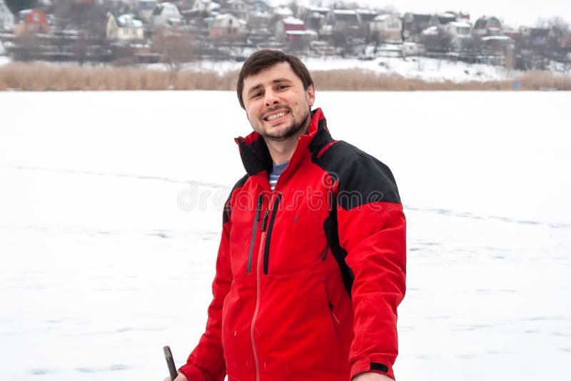A man in a red jacket is smiling, standing on a frozen river. Far beyond the river small houses can be seen.