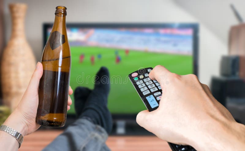 Man is relaxing with legs on table and is watching football match on tv with bear.