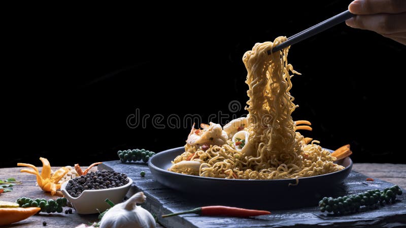 Man`s hand using chopsticks to pick up stir fry instant noodles spicy seafood in black plate with various spices on black slate i