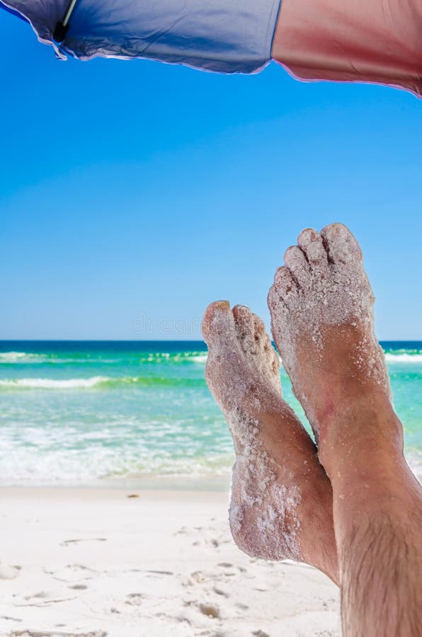 Man`s sandy feet under umbrella, with ocean beach waters in the background