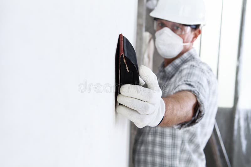 Man sand the wall with sandpaper, professional construction worker with mask, safety hard hat, gloves and protective glasses.